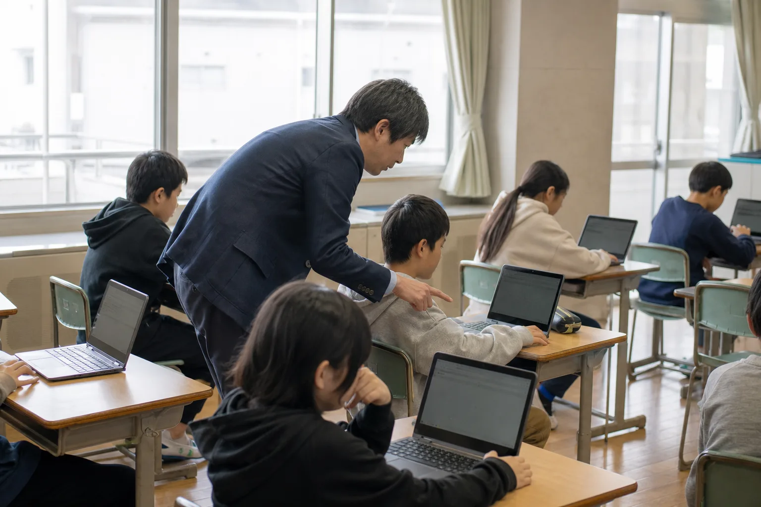 講師が机のあいだを回り、生徒のノートパソコンの画面を指して教えているようす